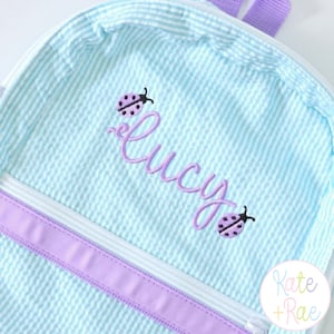 May include: A light blue and white seersucker backpack featuring a purple embroidered name Lucy in cursive script. The design includes two small purple ladybug accents above and below the name, with a purple zipper trim and purple straps.
