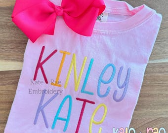 Op de afbeelding: Roze T-shirt met de namen "Kinley" en "Kate" geborduurd in regenboogkleuren. De tekst "Kate + Rae Embroidery" is ook zichtbaar.