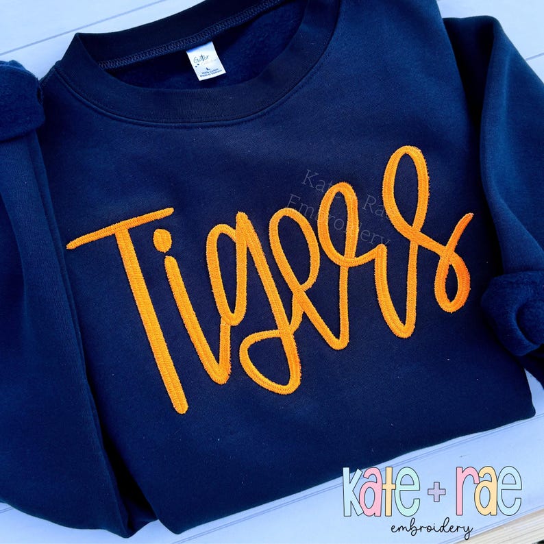 Puede incluir: Sudadera azul marino con la palabra "Tigers" bordada en naranja en la parte delantera. La sudadera tiene cuello redondo y mangas largas. El nombre de la marca "kate + rae embroidery" est&aacute; impreso en la parte inferior.