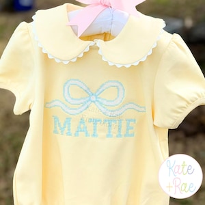 May include: A yellow baby romper with a Peter Pan collar and short sleeves. The romper features a light blue embroidered bow and the name "MATTIE". A pink bow is attached to the hanger.