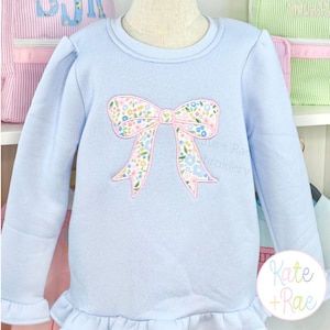 Peut inclure: Un sweat-shirt bleu clair à manches longues avec une applique centrale en forme de nœud ornée d'un motif floral dans des tons de rose, bleu, jaune et vert. Le vêtement présente un col rond côtelé et un ourlet à volants au bas du torse.