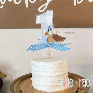 May include: A white frosted cake with a blue and white gingham number one topper and a blue and brown duck topper. The cake is on a gold cake stand. The text "Kate + Rae Embroidery" is visible in the background.