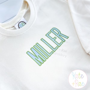 May include: Cream-colored sweatshirt with the name "MILLER" embroidered in light blue with a green outline. The sweatshirt has a crew neck and a small tag on the collar. The brand name "Kate + Rae Embroidery" is also visible.