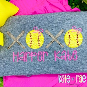 May include: A gray t-shirt with pink embroidery of two baseballs with bows, two crossed bats, and the text "Harper Kate".