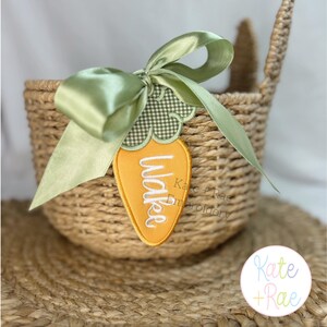 May include: A woven basket with a green ribbon and a carrot-shaped tag. The tag is orange with the name "Wake" embroidered in white. The tag is attached to the basket with a green gingham fabric and a green satin ribbon. The basket is made of natural fibers.