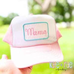 May include: A pink and white trucker hat with the word "Mama" embroidered in pink script on a white patch with a blue border. The hat has a pink brim and mesh back. The text "Kate + Rae Embroidery" is also visible.