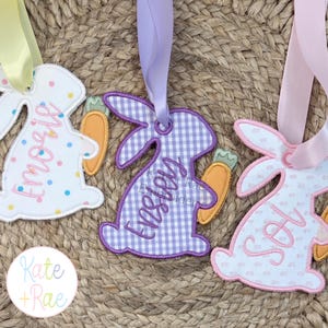 May include: Three Easter bunny-shaped tags with personalized names and carrot accents. One bunny is white with colourful polka dots, another is purple gingham, and the third is white with a pink border. Each has a ribbon for hanging.