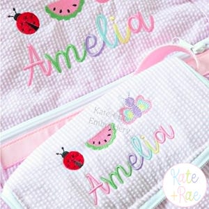 Puede incluir: Un art&iacute;culo de tela seersucker en color lila claro y blanco con dise&ntilde;os bordados de una mariquita roja, una rodaja de sand&iacute;a rosa y una mariposa colorida. El nombre Amelia est&aacute; bordado en una fuente cursiva multicolor sobre la superficie.