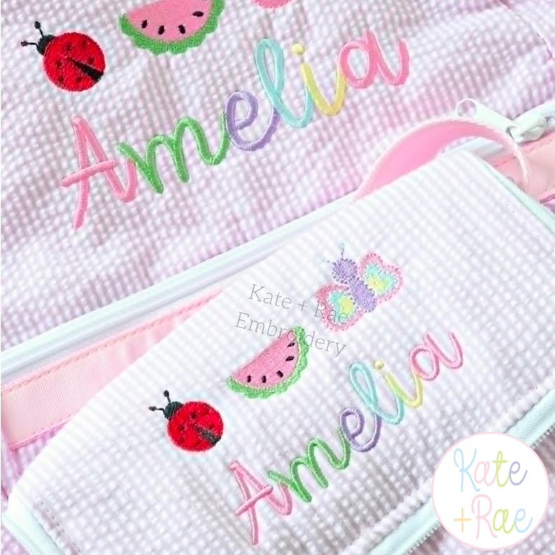 Puede incluir: Un art&iacute;culo de tela seersucker en color lila claro y blanco con dise&ntilde;os bordados de una mariquita roja, una rodaja de sand&iacute;a rosa y una mariposa colorida. El nombre Amelia est&aacute; bordado en una fuente cursiva multicolor sobre la superficie.