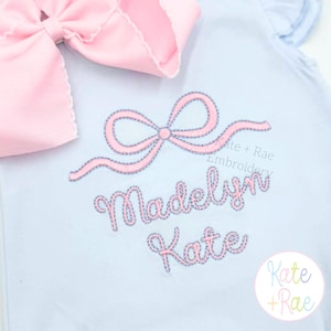 May include: Light blue baby outfit with a large pink bow and embroidered pink bow design. The name "Madelyn Kate" is embroidered in pink script. The brand name "Kate + Rae" is also visible.