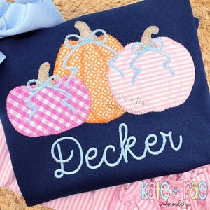 May include: Navy blue long-sleeve shirt with three embroidered pumpkins in orange, pink, and pink gingham. Each pumpkin has a light blue bow. The name "Decker" is embroidered in light blue script. The shirt is from "Kate + Rae embroidery".