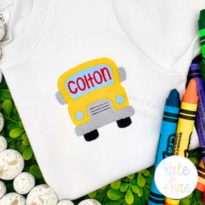 May include: A white t-shirt featuring an embroidered yellow school bus design with the name Colton stitched in red letters across the center. The bus has gray accents and blue headlights. A blue crayon and an orange crayon are positioned nearby.