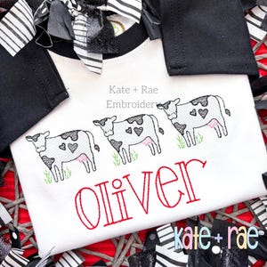 Peut inclure: Combinaison blanche pour bébé avec broderie de vache noire et blanche et le nom "Oliver" en broderie rouge. La combinaison a une bordure rayée noire et blanche autour du cou.