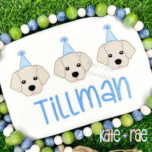 May include: White embroidered shirt with three yellow dogs wearing blue party hats. The shirt says "Tillman" in blue embroidery.