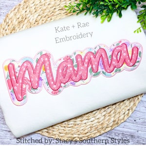 Puede incluir: Bordado rosa de "Mama" con un diseño floral, cosido por Kate + Rae Embroidery. El bordado está sobre un fondo de tela blanca.