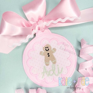 Puede incluir: Un adorno navideño rosa bordado con un diseño de hombre de jengibre y el nombre "Adi" en escritura verde. El adorno está adornado con un lazo rosa y el texto "Kate + Rae Embroidery".