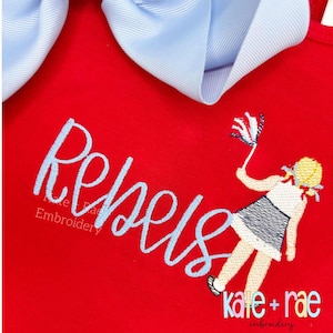 May include: Red garment with the word "Rebels" embroidered in light blue script. A cartoon figure of a girl waving a small flag is also embroidered. A large light blue bow is at the top. The text "kate + rae embroidery" is at the bottom.