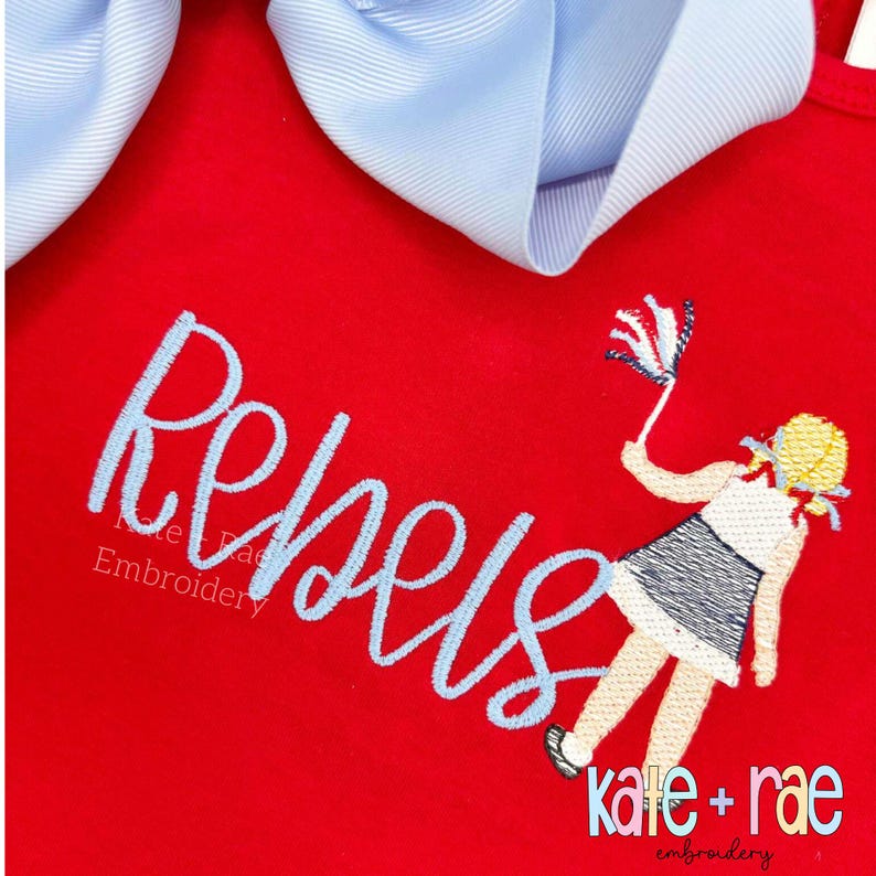 May include: Red garment with the word "Rebels" embroidered in light blue script. A cartoon figure of a girl waving a small flag is also embroidered. A large light blue bow is at the top. The text "kate + rae embroidery" is at the bottom.