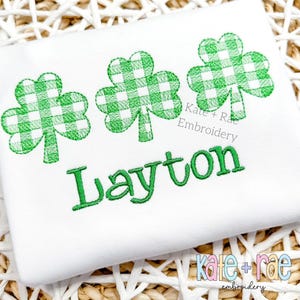May include: White onesie with green embroidered shamrocks and the name Layton. The shamrocks are a checkered pattern. The text "Kate + Rae Embroidery" is also embroidered on the onesie.