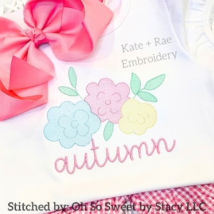 Puede incluir: Una camiseta blanca con un dise&ntilde;o floral bordado en azul claro, rosa y amarillo con hojas verdes. El nombre autumn est&aacute; bordado en cursiva rosa debajo de las flores. Un lazo rosa grande est&aacute; sujeto y el texto Kate + Rae Embroidery es visible.