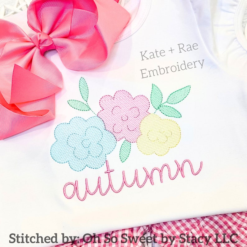 Puede incluir: Una camiseta blanca con un dise&ntilde;o floral bordado en azul claro, rosa y amarillo con hojas verdes. El nombre autumn est&aacute; bordado en cursiva rosa debajo de las flores. Un lazo rosa grande est&aacute; sujeto y el texto Kate + Rae Embroidery es visible.