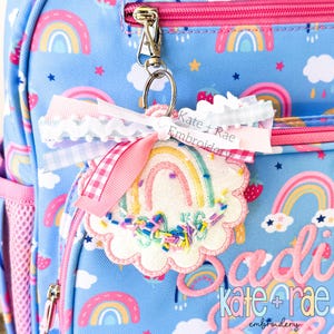 May include: A blue backpack with a rainbow design, featuring a rainbow-shaped keychain. The keychain has a rainbow design with colourful sprinkles and the text "Kate + Rae Embroidery". The keychain is attached to the backpack with a silver clasp.