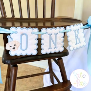 May include: A white and light blue "ONE" banner with a scalloped edge is displayed on a wooden high chair. The banner features the word "ONE" in light blue letters, with a puppy wearing a party hat on the "O". The banner is tied with light blue ribbon.