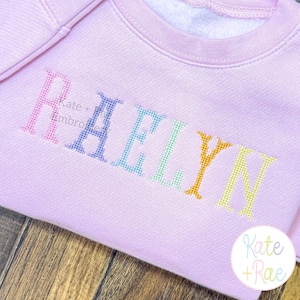 May include: A light pink sweatshirt with the name "RAELYN" embroidered in a rainbow of colors. The sweatshirt has a crew neck and the brand name "Kate + Rae" is visible in the image.