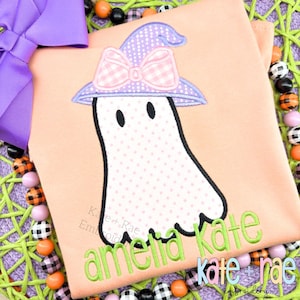 Puede incluir: Camiseta color melocotón con un fantasma bordado que lleva un sombrero de bruja y un lazo rosa. El fantasma tiene un estampado de lunares. El nombre "amelia kate" está bordado en verde. La marca "Kate + Rae Embroidery" también es visible.