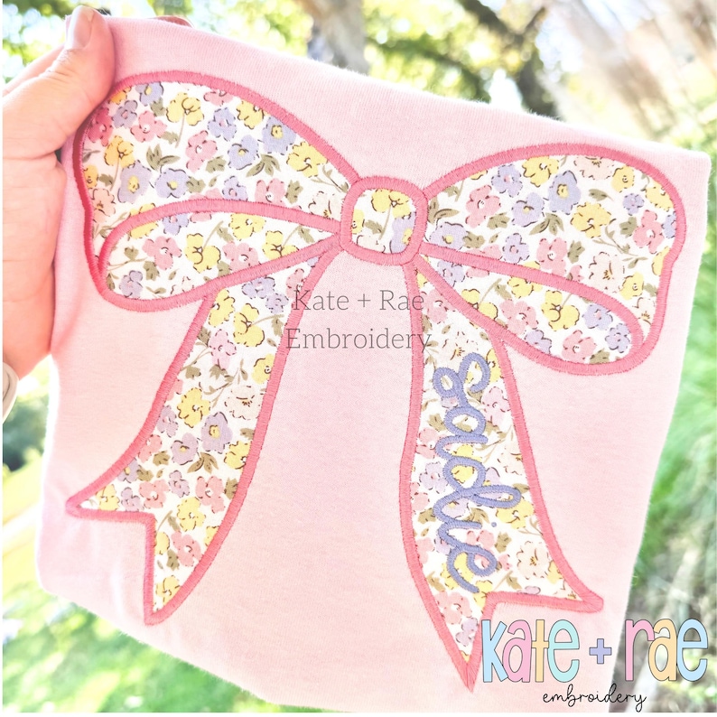 Puede incluir: Una camiseta rosa claro con una gran aplicaci&oacute;n de lazo bordado con un estampado floral en tonos amarillo, morado y verde. El nombre Sadie est&aacute; bordado en cursiva morada en uno de los extremos del lazo. El texto dice Kate + Rae Embroidery.