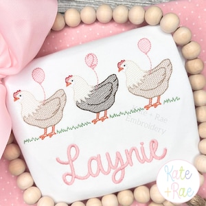 May include: White shirt embroidered with three chickens holding pink balloons. The name "Laynie" is embroidered in pink script below the chickens. The design includes green grass and the text "Kate + Rae Embroidery".