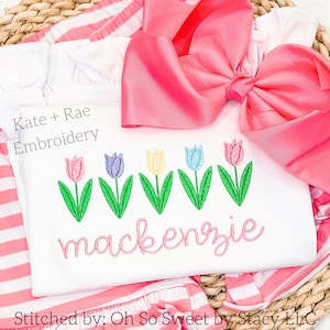 May include: White embroidered shirt with a row of five colorful tulips and the name "mackenzje" in pink script. The shirt is on a woven straw surface with a pink bow.