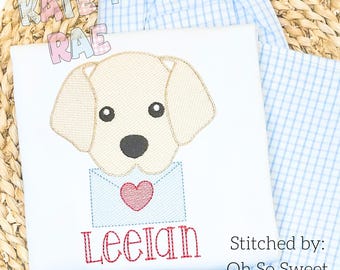Op de afbeelding: Een wit T-shirt met een geborduurd beeld van een hond die een blauwe envelop met een rood hart vasthoudt. De hond is beige met zwarte ogen en neus. De naam "Leeian" is in rood onder de hond geborduurd. De tekst "Stitched by: Oh So Sweet by Stacy LLC" is in zwart in de rechterbenedenhoek van het T-shirt geborduurd.
