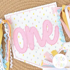 May include: A white fabric pennant with scalloped edges featuring the word one in large pink script lettering. The background fabric has a pattern of small yellow and blue dots. The pennant is decorated with various pastel ribbons on the sides.
