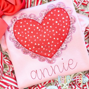 May include: Pink shirt with a large heart-shaped design featuring a red heart with smaller pink and white hearts inside. The heart is surrounded by a pink and white sequined border. The name "annie" is embroidered below the heart in pink thread. The text "kate + rae embroidery" is also embroidered in pink and white thread at the bottom of the shirt.