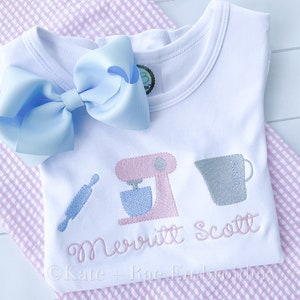 May include: White baby onesie with pink, blue, and gray embroidery of a rolling pin, a stand mixer, and a measuring cup. The onesie is personalized with the name "Merritt Scott".