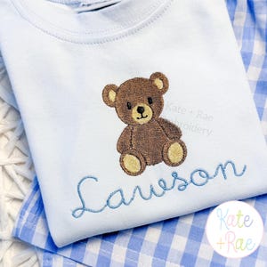May include: A light blue baby shirt with an embroidered brown teddy bear and the name "Lawson" in blue script. The shirt is folded on a blue and white gingham patterned fabric. The brand name "Kate + Rae" is also visible.