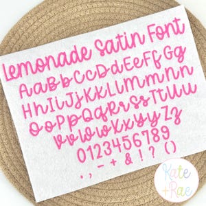May include: A white sheet featuring the Lemonade Satin Font in bright pink. The display includes uppercase and lowercase letters from A to Z, numbers 0 through 9, and symbols including an ampersand, exclamation point, question mark, and parentheses.