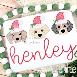 May include: White embroidered shirt with three dogs wearing Santa hats. The dogs are brown, black and white, and yellow. The name "henley" is embroidered in red below the dogs. The text "kate + rae embroidery" is at the bottom of the shirt.