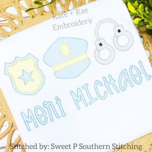 May include: A white embroidered design with a blue police hat, a yellow star badge, and handcuffs. The name "Michael" is embroidered in blue below the design. The text "Kate + Rae Embroidery" is at the top of the design. The text "Stitched by: Sweet P Southern Stitching" is at the bottom of the design.