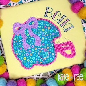 May include: A yellow fabric square with an embroidered design. The design features a blue floral pattern with a purple bow and the name "Bella" in a decorative font. The edges are trimmed with pink stitching.