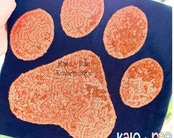 May include: A navy blue fabric with a large paw print design. The paw print is made of orange sequins. The text "Kate + Rae Embroidery" is visible in the image.