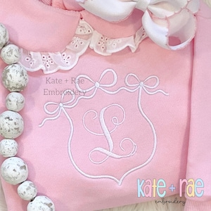 May include: Pink sweatshirt with a white embroidered monogram and bows. The text "Kate + Rae Embroidery" is also embroidered on the sweatshirt.