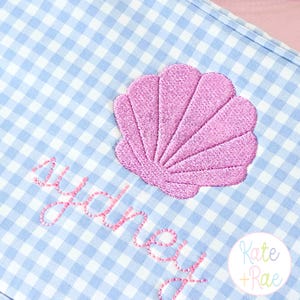 May include: A light blue and white gingham pattern fabric with a pink embroidered seashell and the name "Sydney" in pink script. The lower right corner features a circular logo with the text "Kate + Rae".