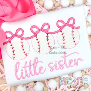 Op de afbeelding: Een wit shirt met roze borduurwerk van drie honkballen met roze strikken en de tekst "little sister". Het shirt heeft ook de tekst "kate + rae embroidery" in roze.