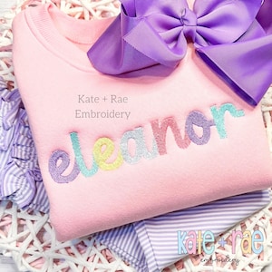 Può includere: Felpa rosa con il nome "Eleanor" ricamato in colori arcobaleno. Il testo "Kate + Rae Embroidery" è visibile anche sulla felpa.