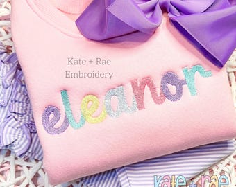 May include: Pink sweatshirt with the name "Eleanor" embroidered in rainbow colors. The text "Kate + Rae Embroidery" is also visible on the sweatshirt.