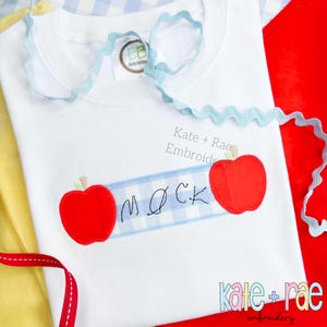 May include: White t-shirt with a light blue ruffled collar and trim. The shirt features two red apple appliqués flanking a blue and white gingham banner with the embroidered word "MOCK". The text "Kate + Rae Embroidery" is also visible.