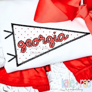 May include: A white shirt with a red bow and a black and white pennant with the word "georgia" in red script. The pennant has a red polka dot pattern.