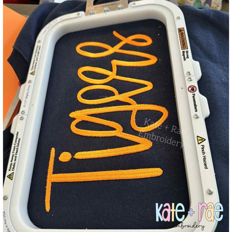 Puede incluir: Primer plano de una tela azul marino con la palabra "Tigers" bordada en hilo naranja brillante. La tela se sujeta en un bastidor de bordado blanco. El texto "Kate + Rae Embroidery" es visible.
